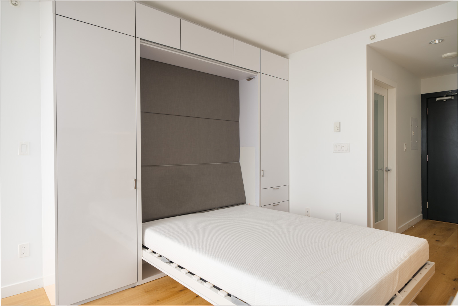 A white Murphy bed is partially unfolded from a built-in wall unit in a minimal, modern room with light wood flooring.