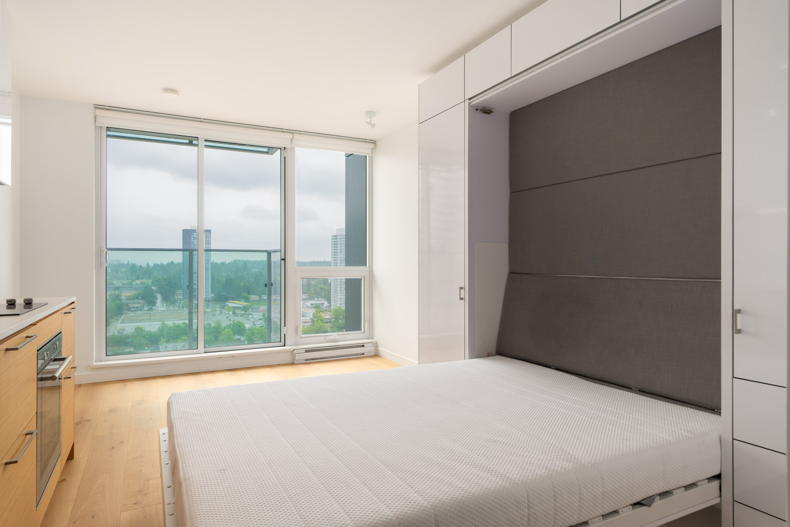 A modern, minimalist apartment with a Murphy bed, large window, light wood flooring, and a small kitchen area, featuring a city view in the background.