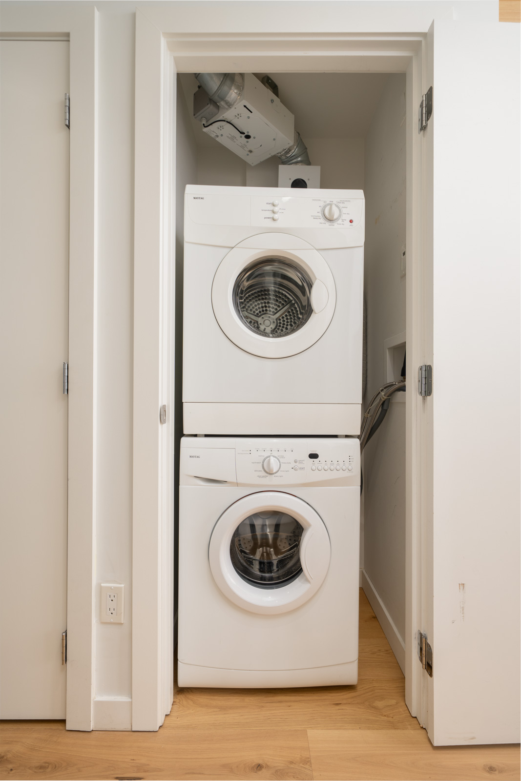 A stacked white washer and dryer are installed in a small closet with white walls and a light wood floor.