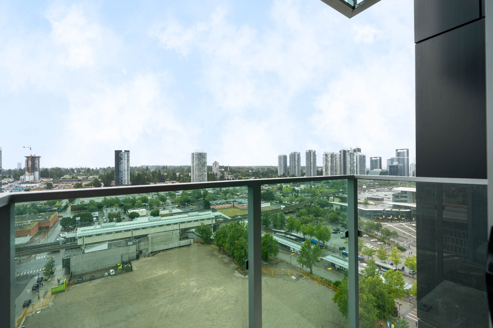 View from a high-rise balcony overlooking a cityscape with various buildings, greenery, and a construction site below, under a partly cloudy sky.