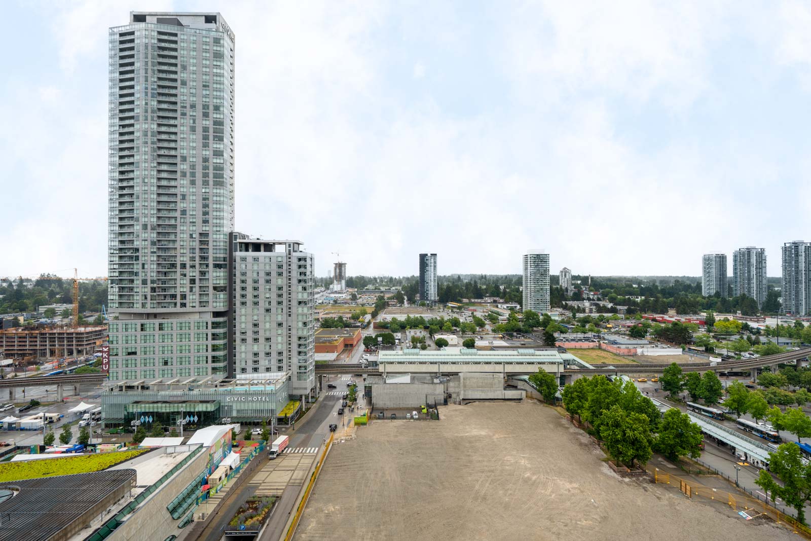 A tall modern high-rise building stands near a vacant lot, with several other high-rise buildings and trees visible in the background under a cloudy sky.