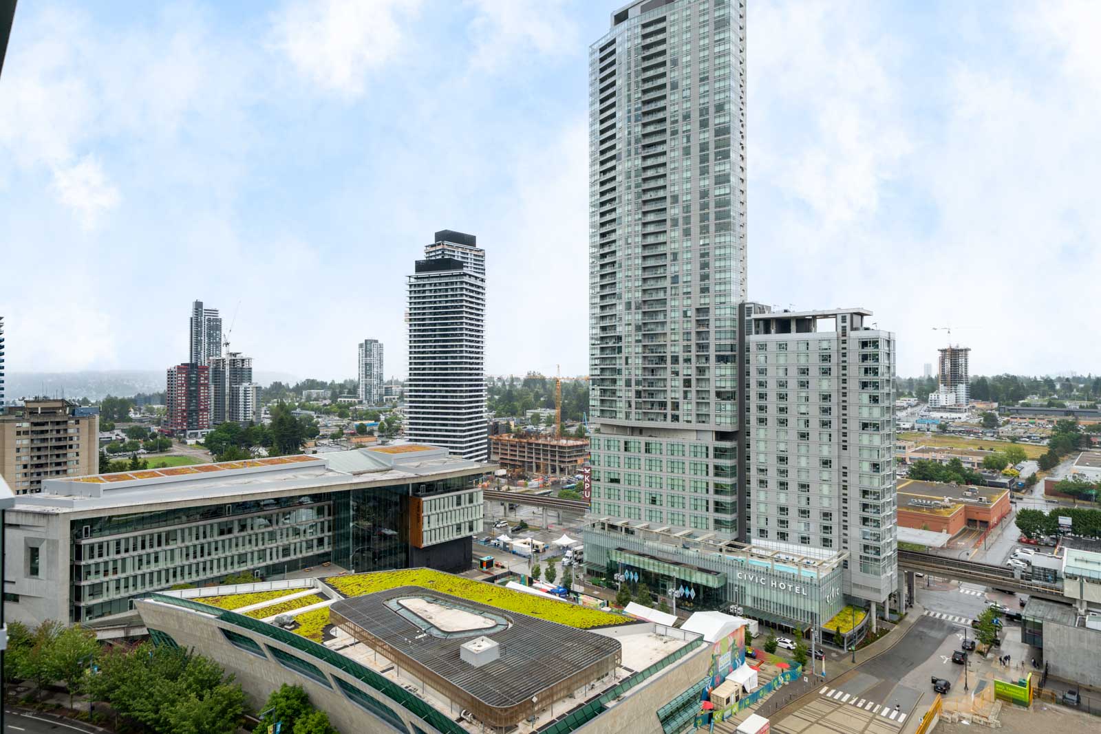 View of a modern cityscape featuring tall residential and commercial buildings, green rooftops, and streets with light traffic under a cloudy sky.