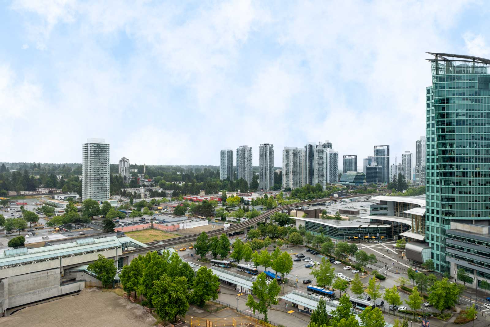 A cityscape with multiple modern high-rise buildings, roads, and green spaces under a partly cloudy sky.