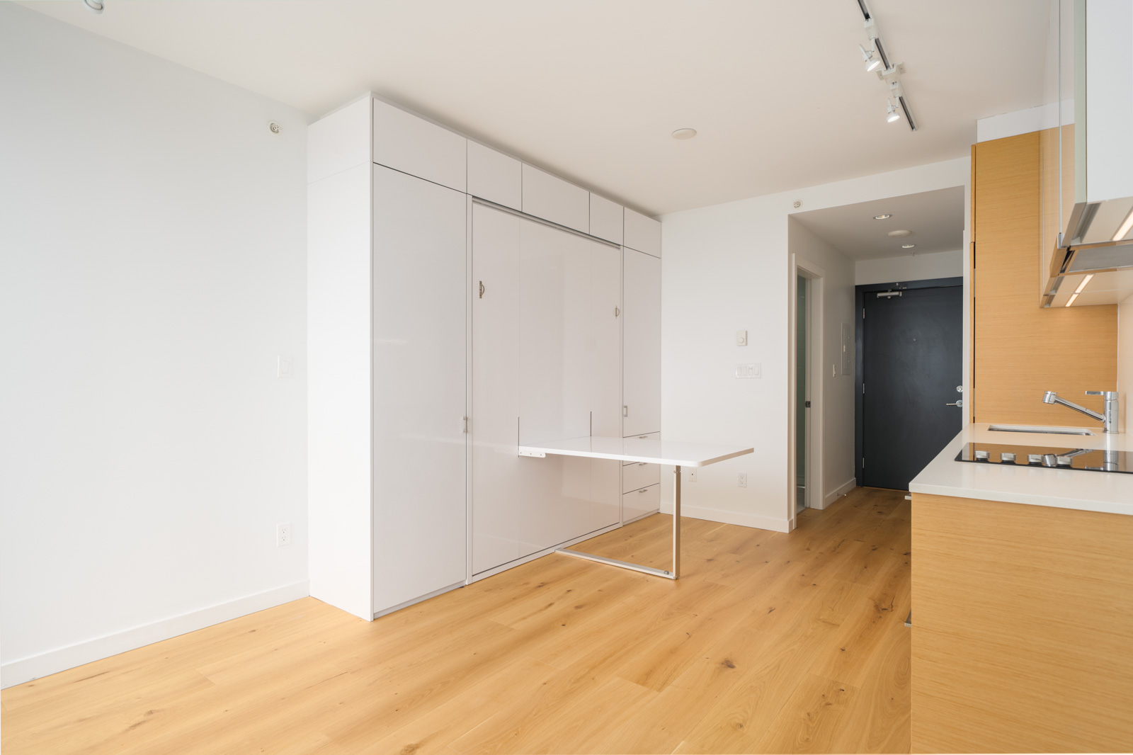 Modern minimalist kitchen with light wood flooring, white cabinets, a fold-down wall table, and built-in appliances. The kitchen opens to a hallway with a dark entry door.