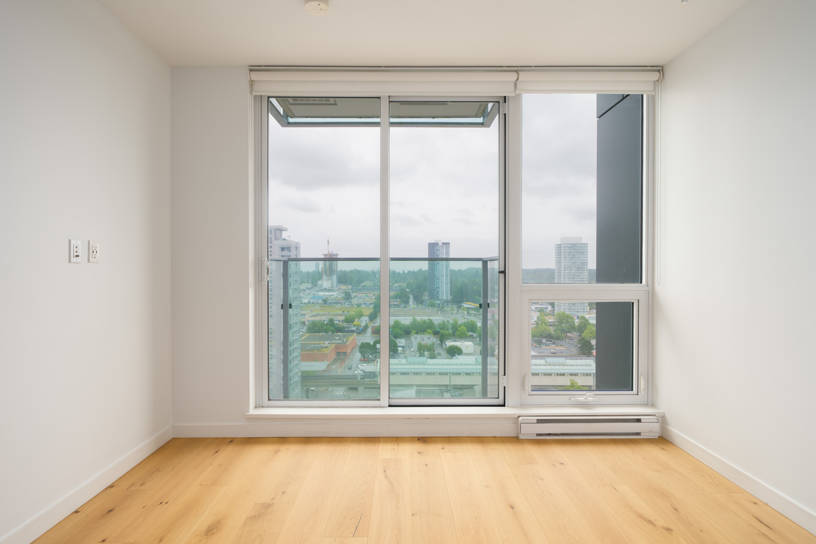 Empty room with light wood flooring, white walls, and large floor-to-ceiling windows overlooking a cityscape with several tall buildings.