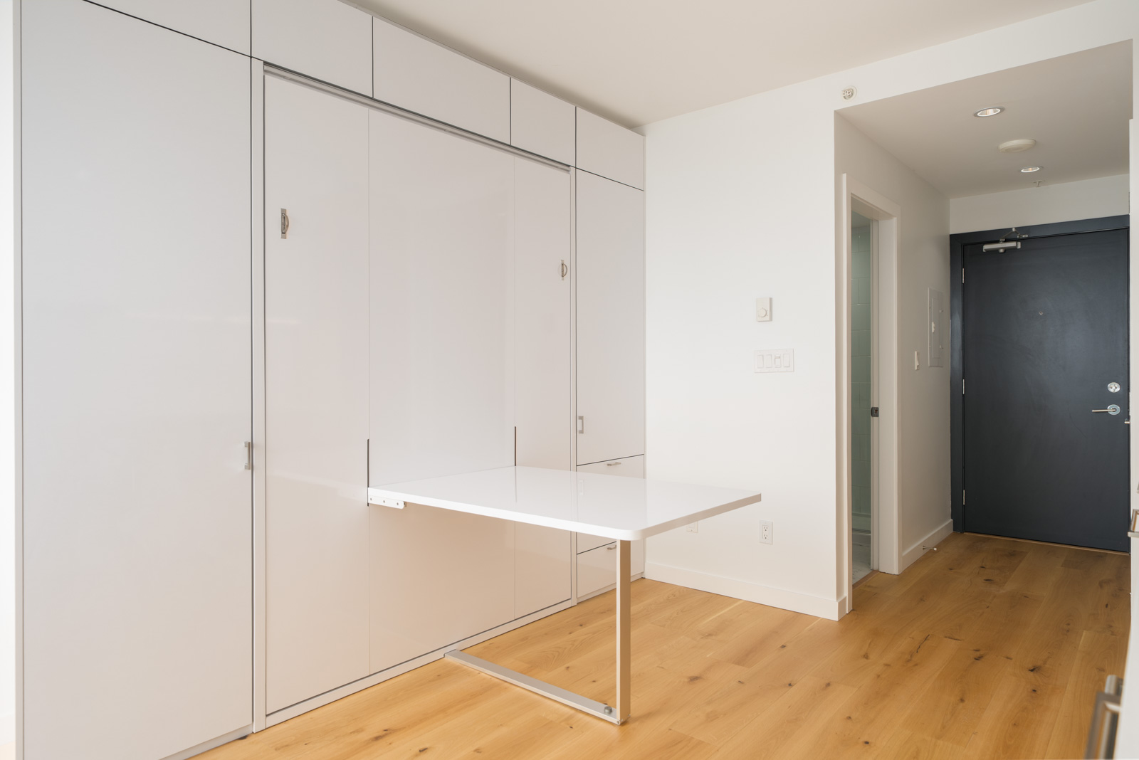 Minimalist room with light wood floor, white walls, built-in white cabinets, a fold-down table, recessed lighting, and a dark entry door.