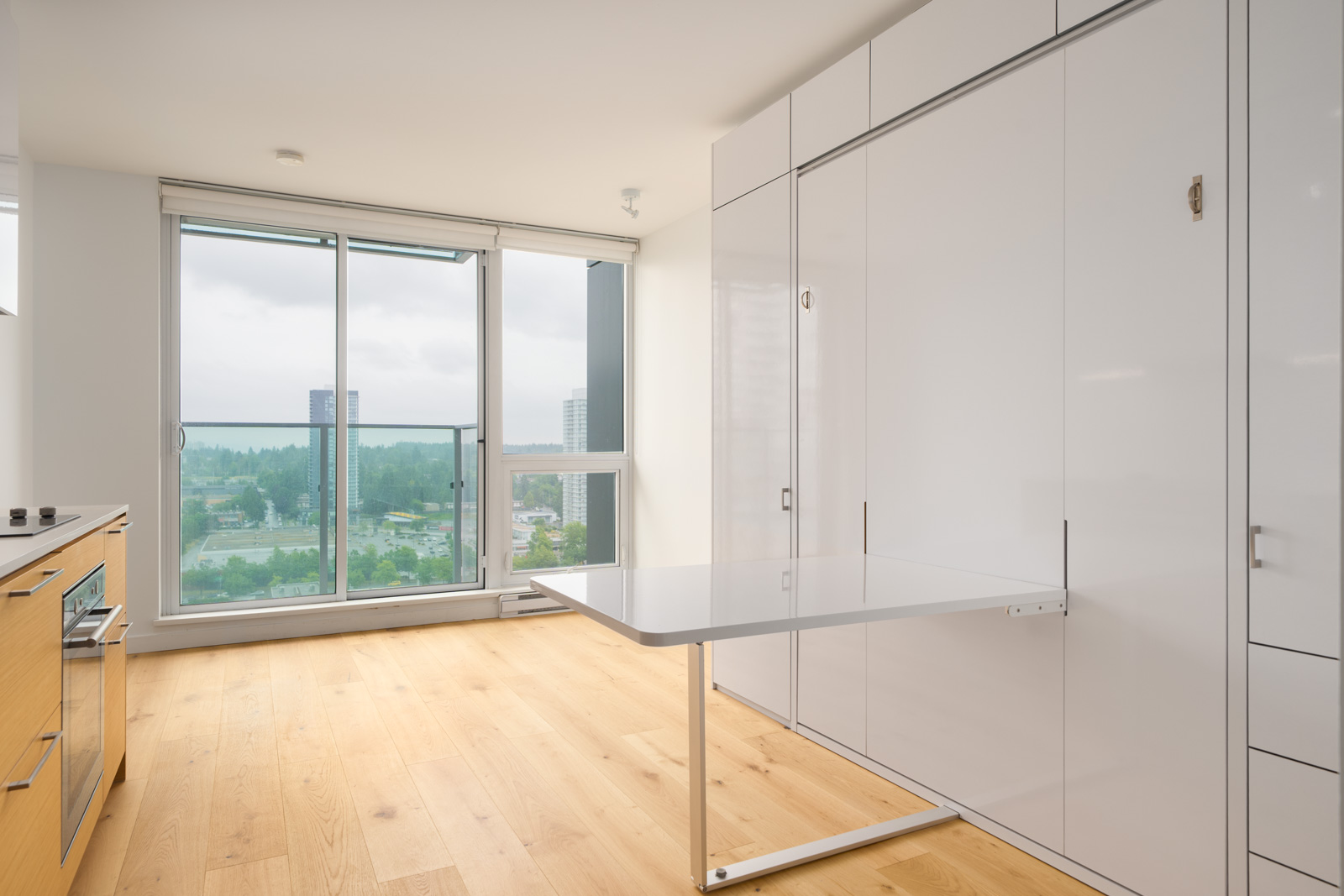 Minimalist apartment interior with light wood flooring, white built-in cabinets, a fold-out table, and large windows overlooking a cityscape and greenery.