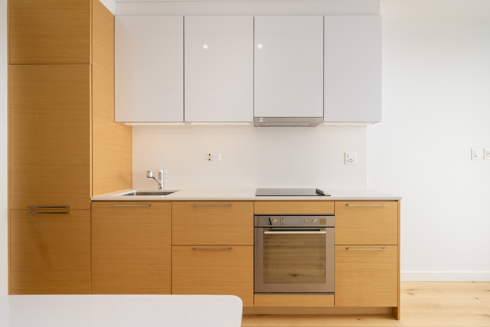 Modern kitchen with light wood cabinets, white upper cabinets, built-in oven, electric cooktop, sink, and minimal decor against a white wall.