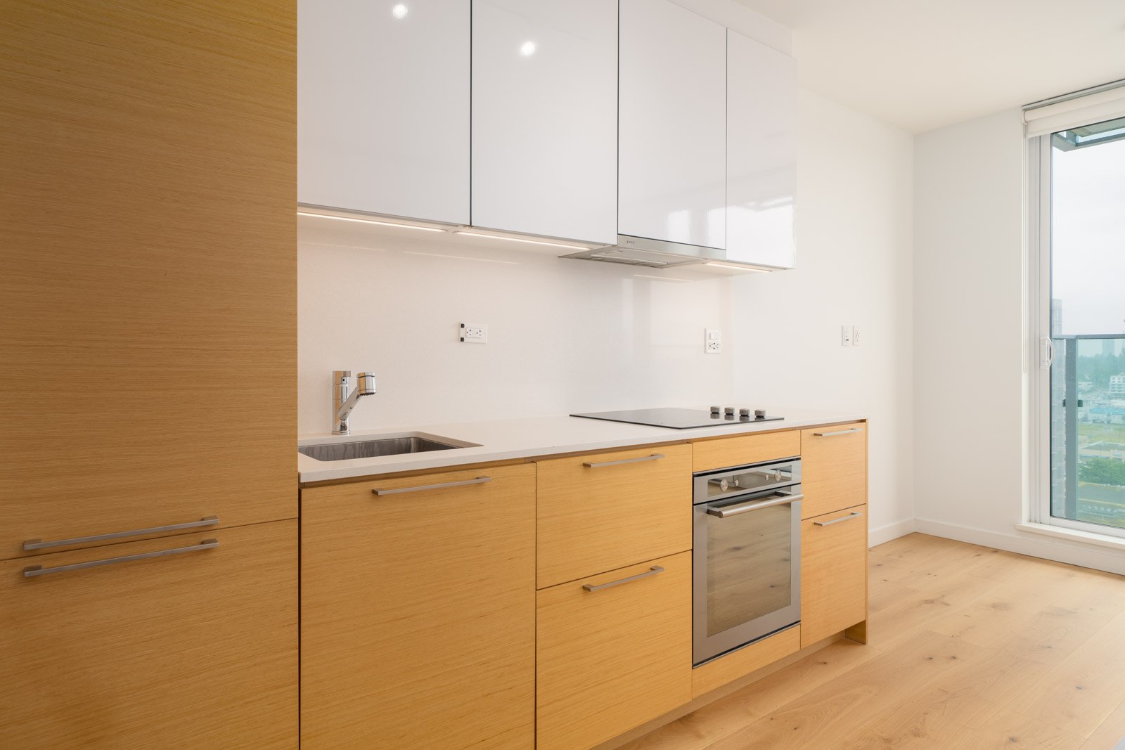Modern kitchen with light wood cabinets, built-in oven, electric stove, white upper cabinets, sink, and large window providing natural light.