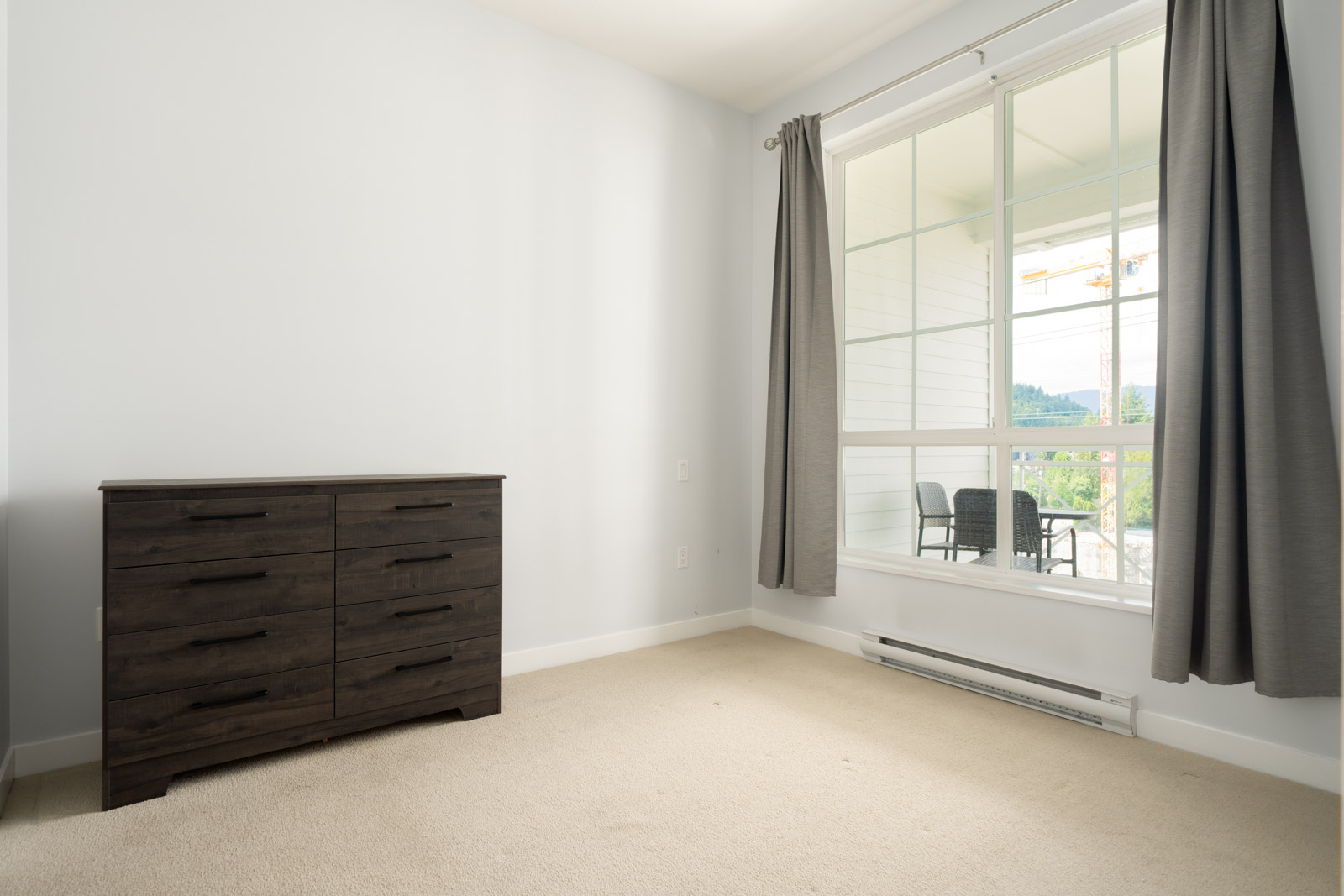 A minimalist bedroom with beige carpet, a dark wooden dresser, grey curtains, and a large window showing a balcony and greenery outside.
