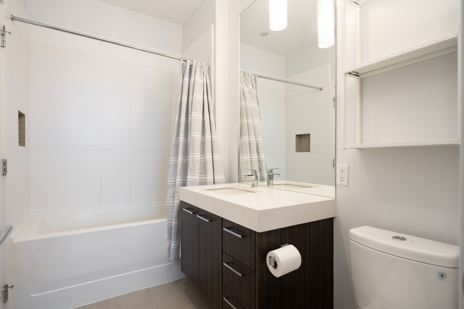Modern bathroom with a bathtub and striped shower curtain, a dark wood vanity with double sinks, large mirror, open shelving, and a white toilet.