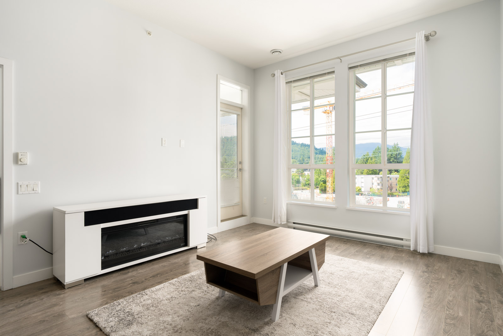 A bright, modern living room with large windows, light-colored walls, a fireplace, and a wooden coffee table on a rug.