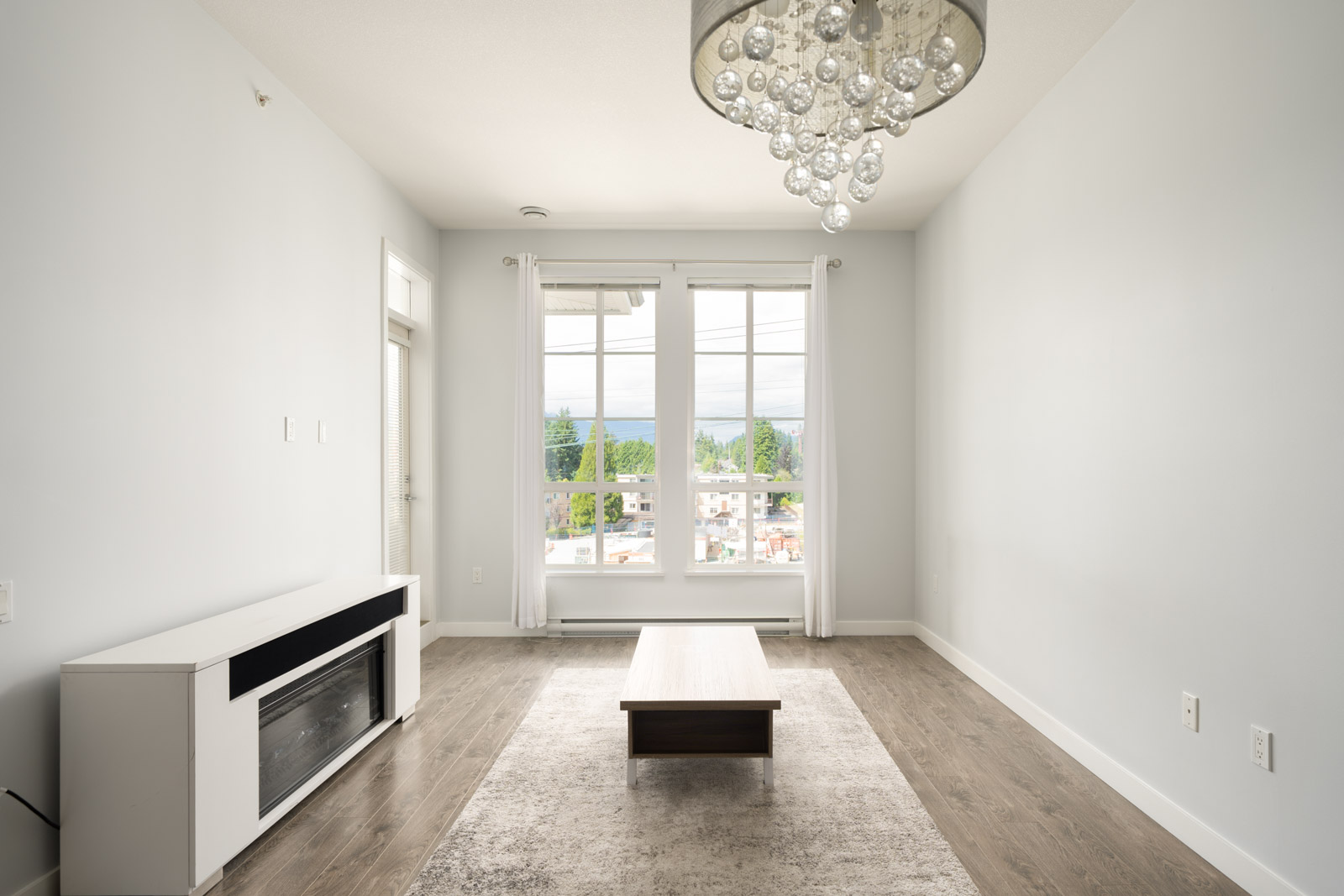 Minimalist living room with a chandelier, white walls, a small coffee table on a rug, a white console, large window, and wood flooring.
