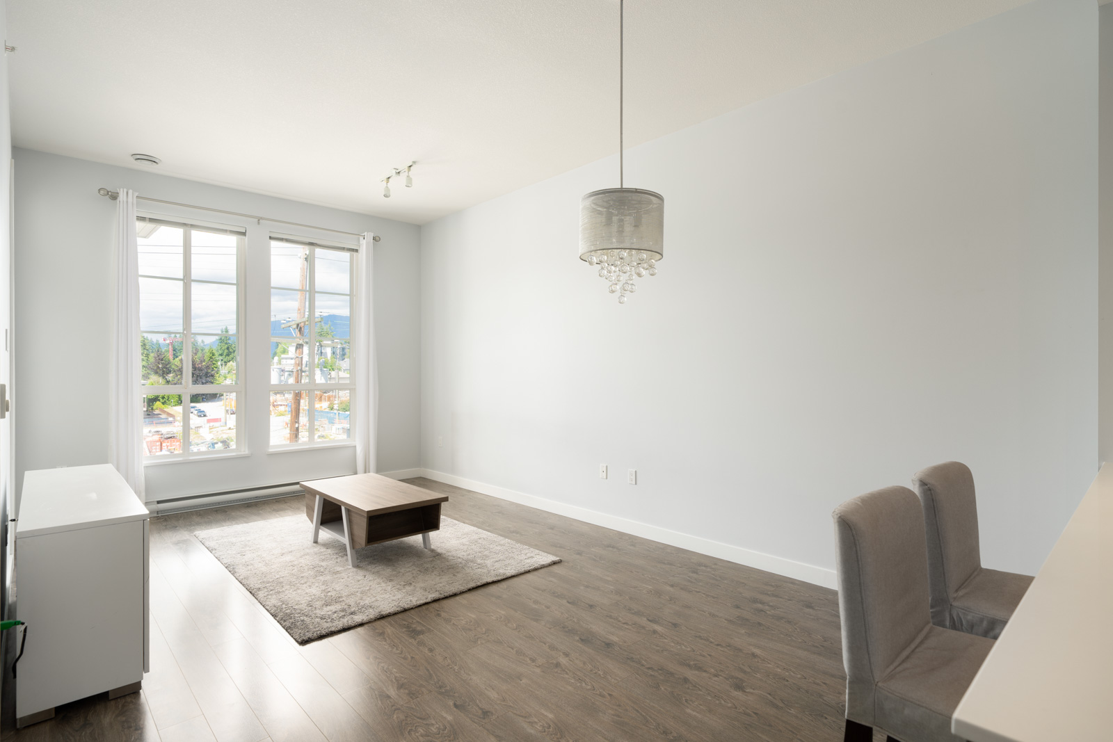 A minimalist living room with light walls, large windows, wood flooring, a small coffee table on a rug, two chairs, and a ceiling light fixture.