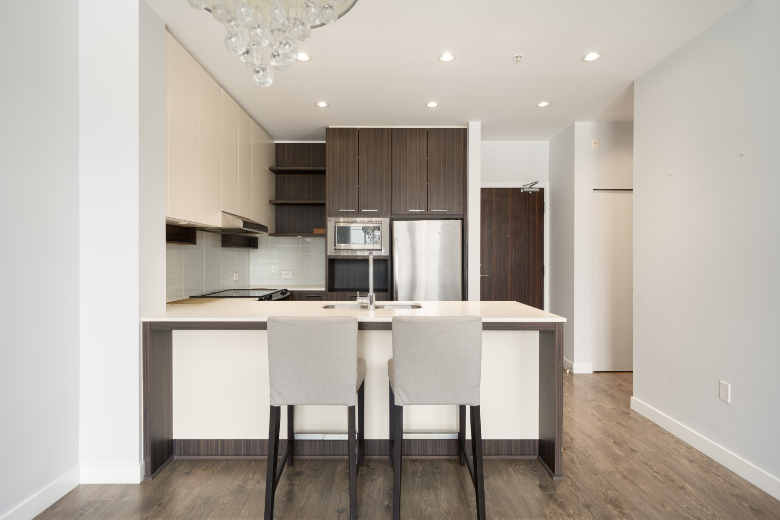 Modern kitchen with white countertops, two beige barstools, stainless steel appliances, dark wood cabinets, and light wood flooring.