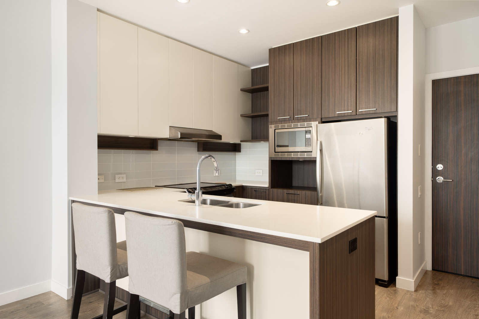 Modern kitchen with wood cabinets, a stainless steel fridge, built-in microwave, electric stove, white countertops, an island with sink, and two upholstered bar stools.