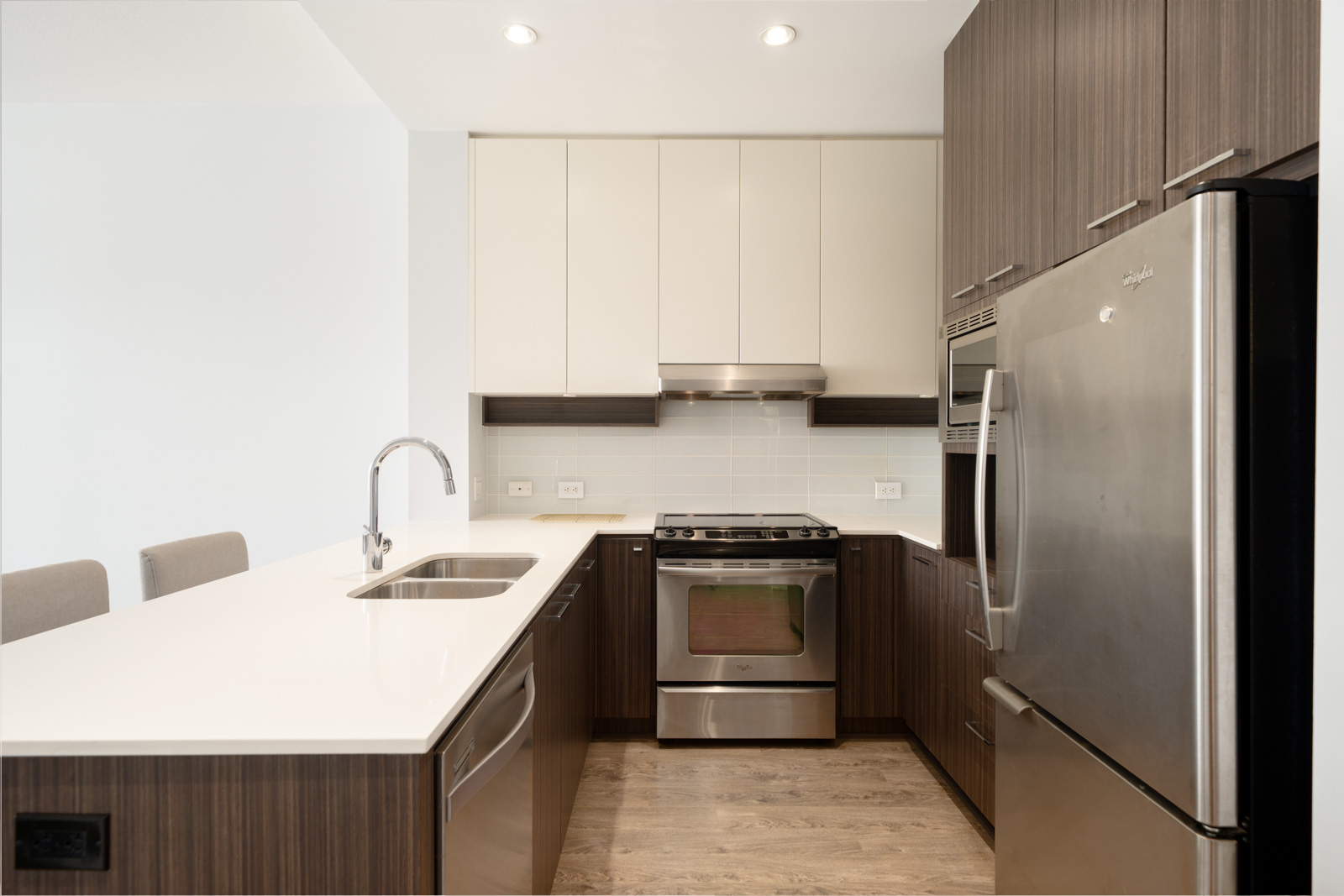 Modern kitchen with wood cabinets, stainless steel appliances, white countertops, and a built-in oven, viewed from the entrance.