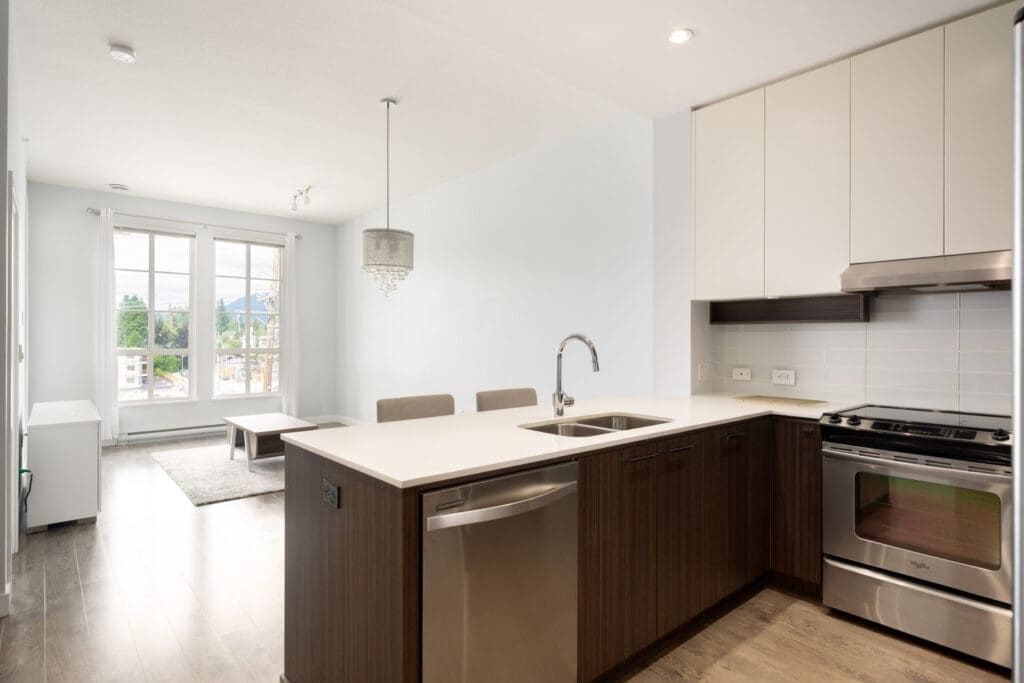 Modern kitchen with stainless steel appliances, white cabinets, and an island with a sink, opening into a bright living room with large windows.
