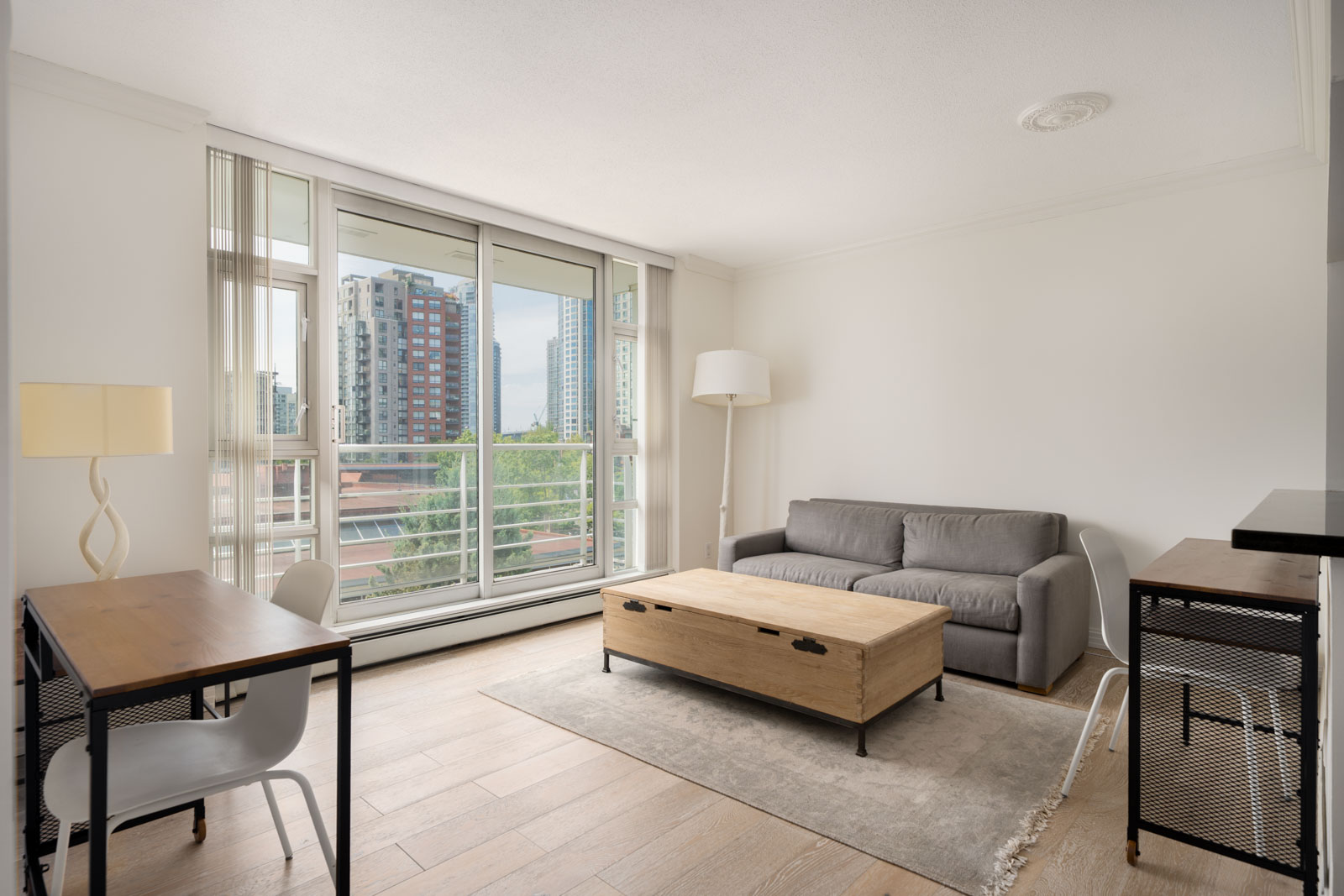 Modern living room with grey sofa, wooden coffee table, two desks, floor lamp, large window, and Vancouver city view.