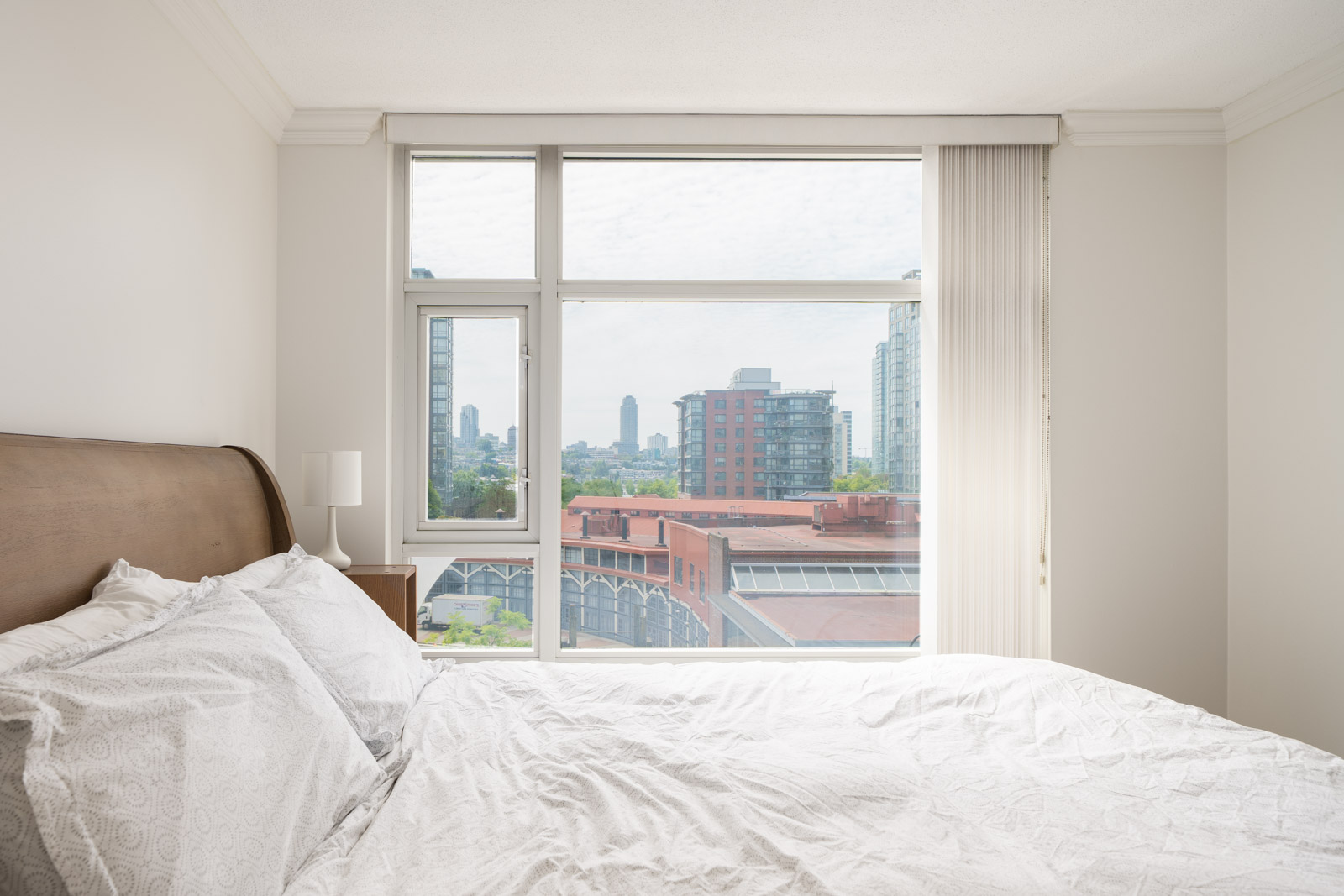 A neatly made bed with white bedding beside a window overlooks city buildings and a partly cloudy sky in Vancouver.