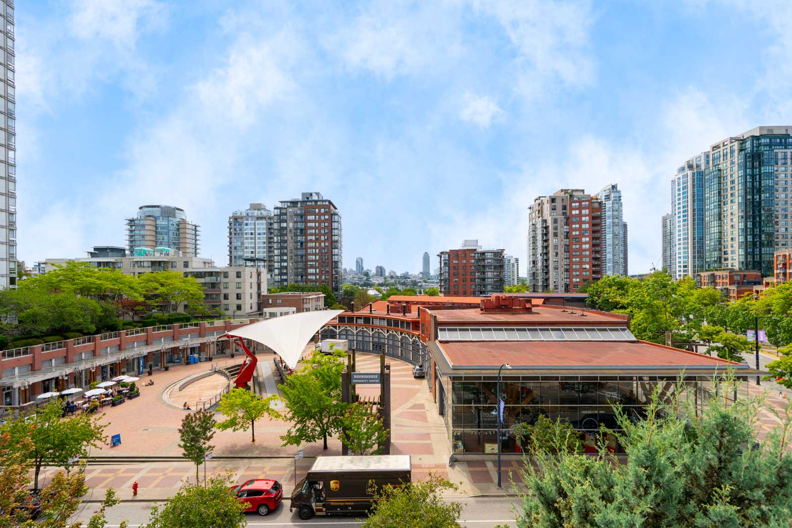 Sunny urban plaza near 189 Davie St, Vancouver, with high-rises, a canopy, glass-roofed building, street trees, and parked cars.