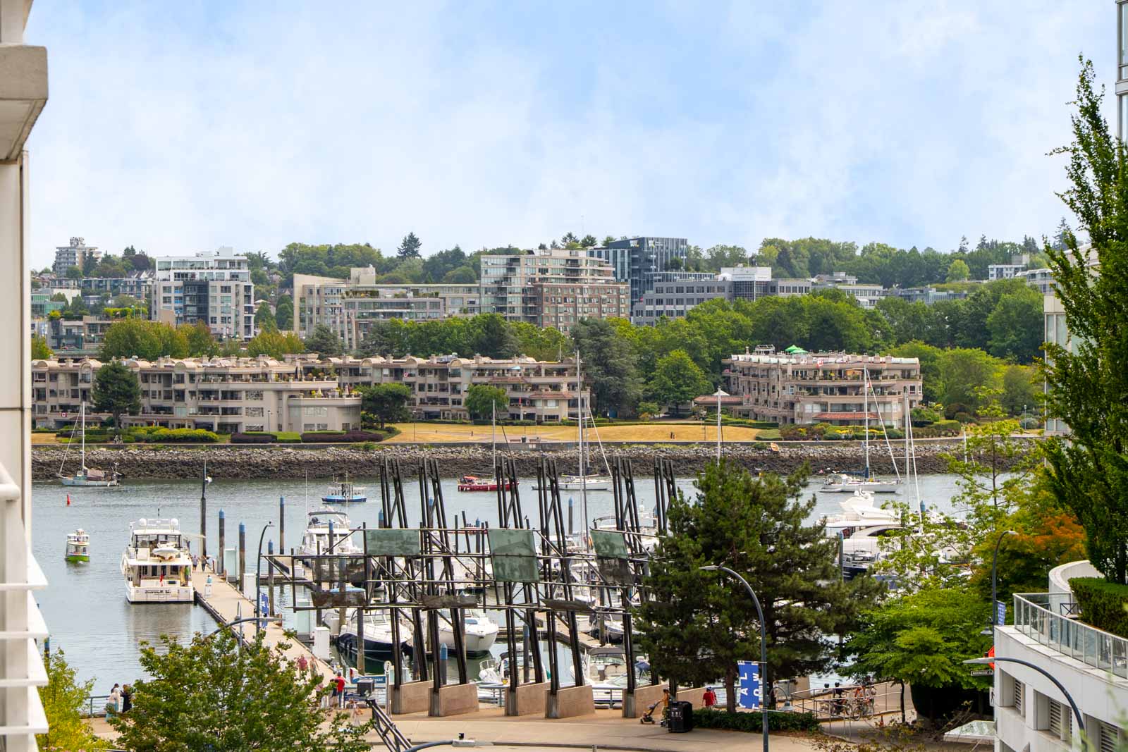 Marina with docked boats in the foreground, trees and 189 Davie St apartment buildings, Vancouver, under partly cloudy skies.
