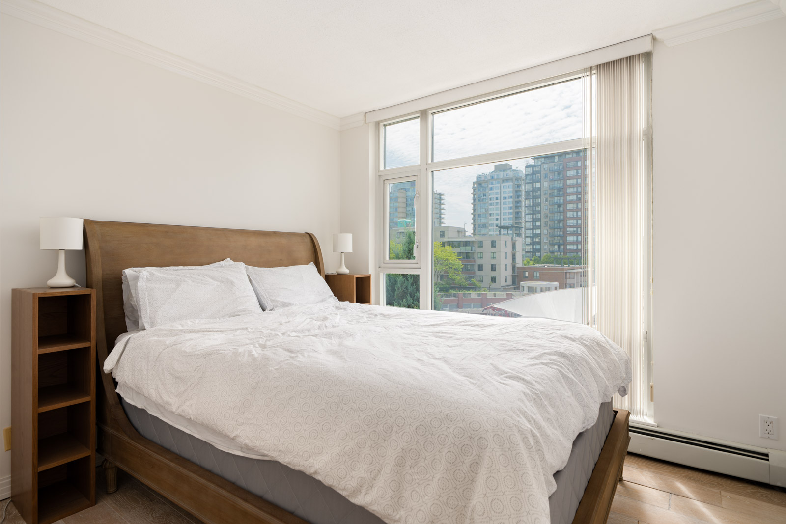 Modern bedroom with wooden bed, white bedding, two bedside tables, large window, and cityscape view at 189 Davie St, Vancouver.