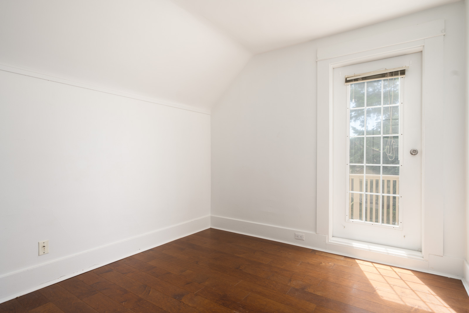 Empty white room with hardwood floors, closed glass door with vertical blinds, sunlight streaming in; Vancouver location.