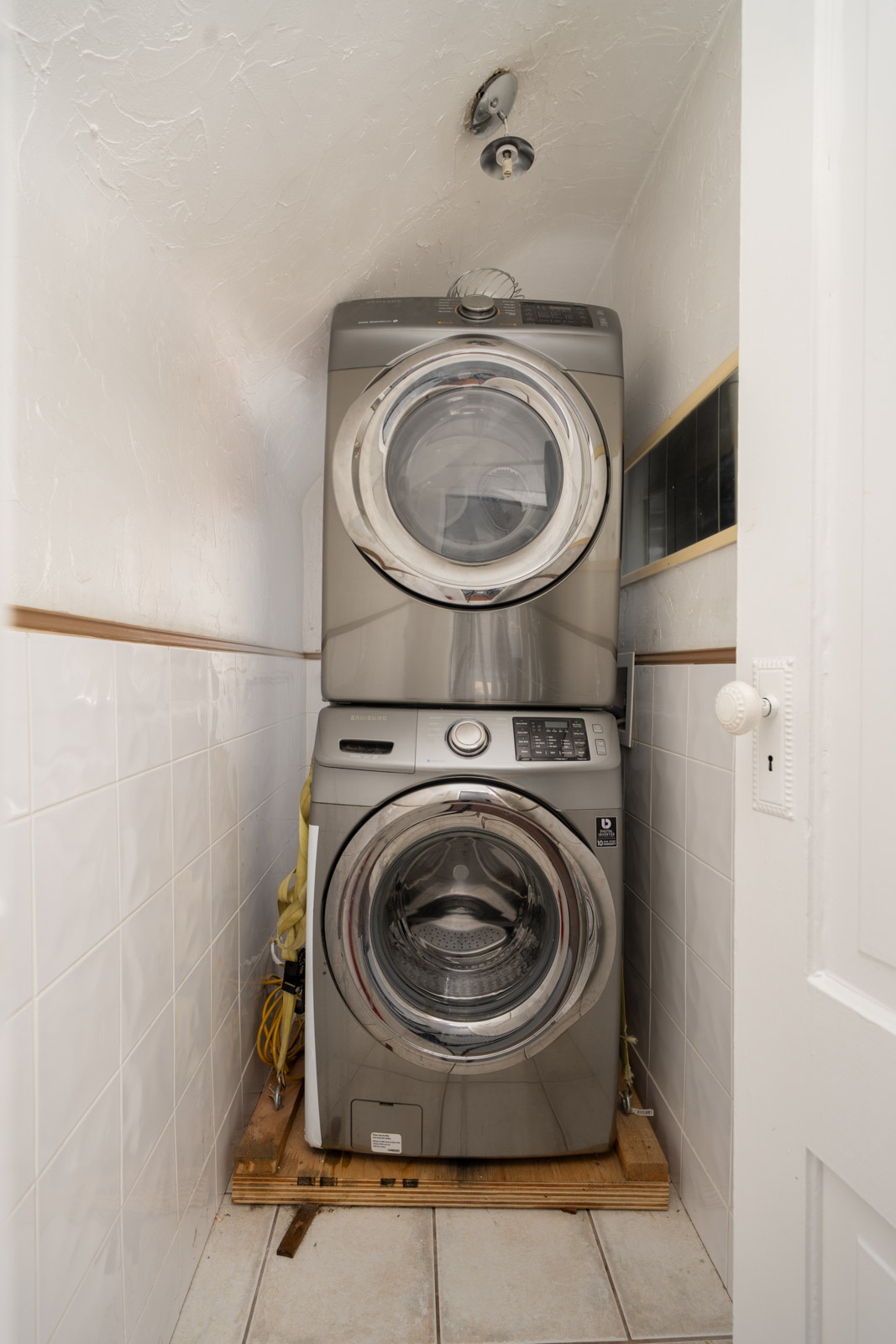 Stacked washer and dryer in a small, tiled laundry nook with a partially open white door in a Vancouver E 19th Ave home.