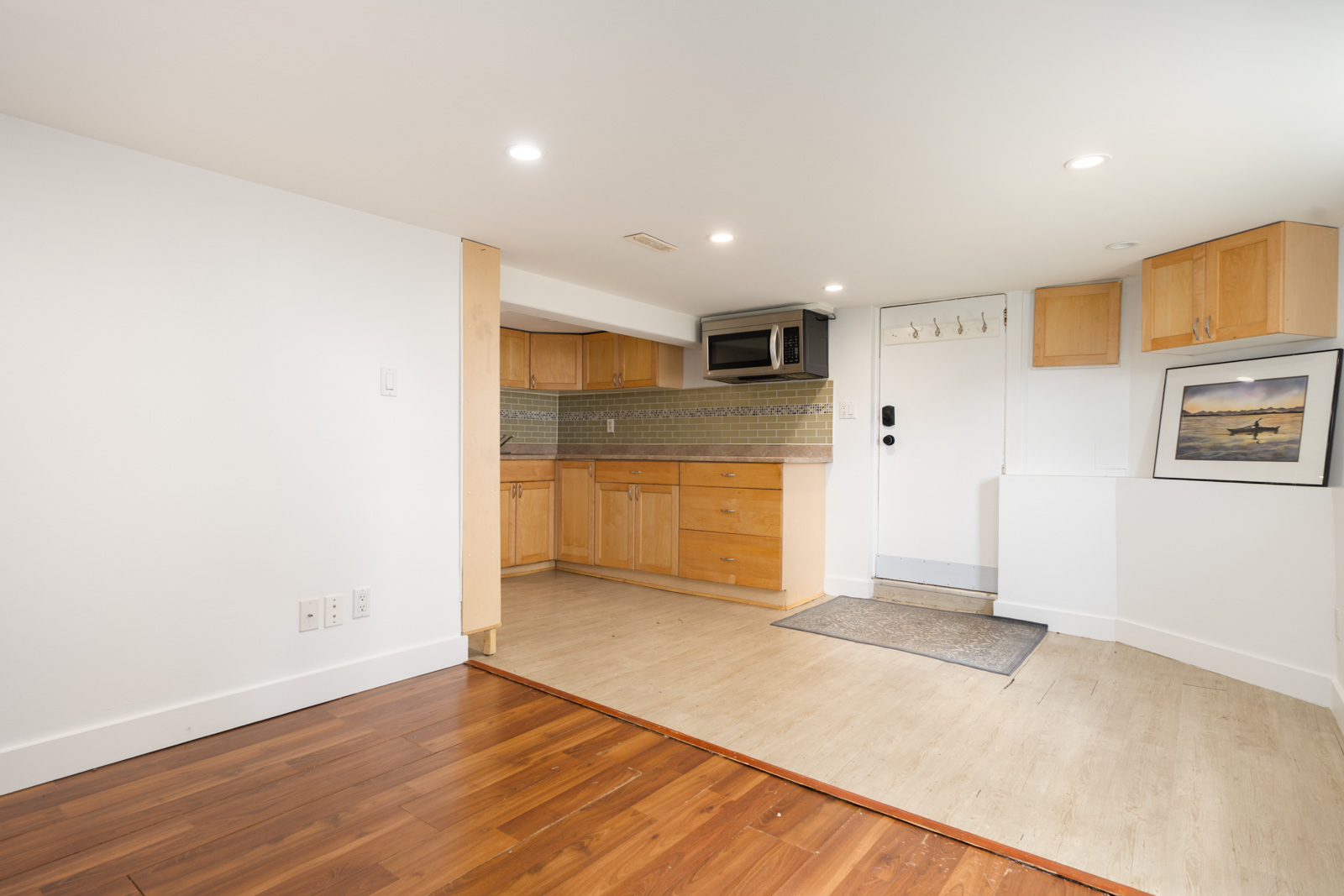 Light wood cabinets in a small Vancouver kitchen by a white entry door; tile floor meets wood, framed photo and recessed lights present.
