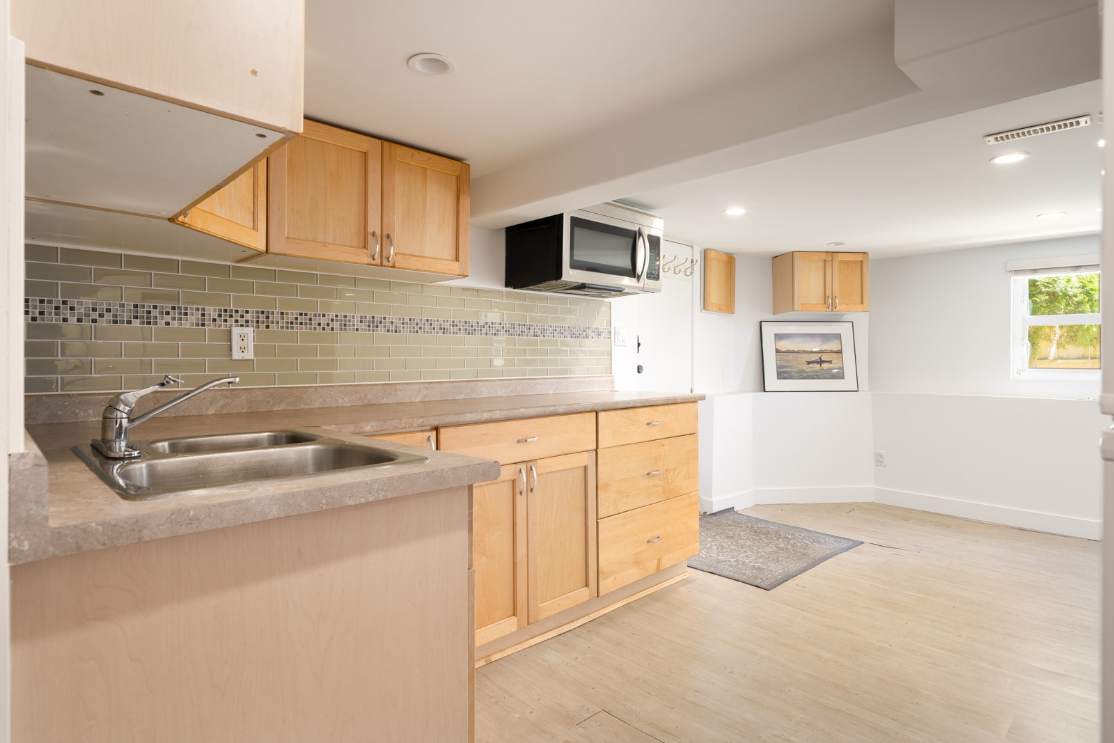 Modern basement kitchen in Vancouver with light wood cabinets, beige counters, sink, microwave, tile backsplash, and small window.