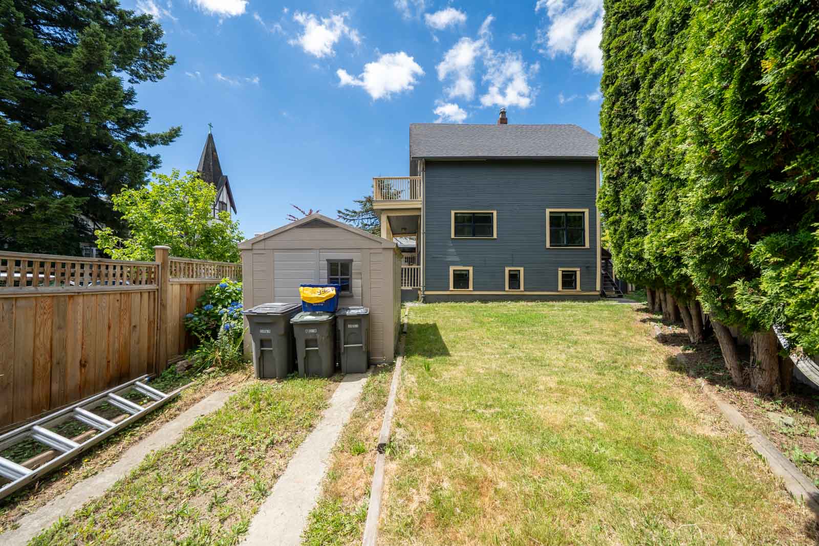 Backyard in Vancouver near E 19th Ave with a gray shed, three trash bins, grass lawn, tall trees on right, and two-story house.