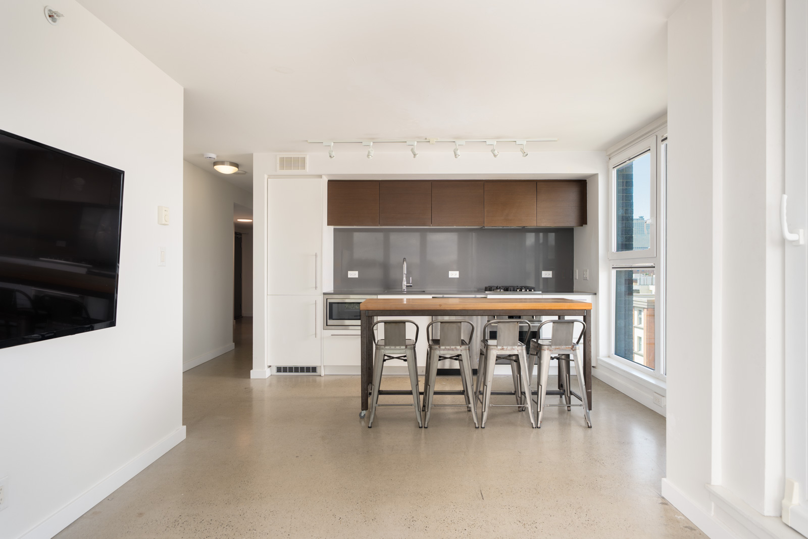 Modern kitchen featuring a wooden island, four metal stools, gray backsplash, white walls, and large sunlit windows.