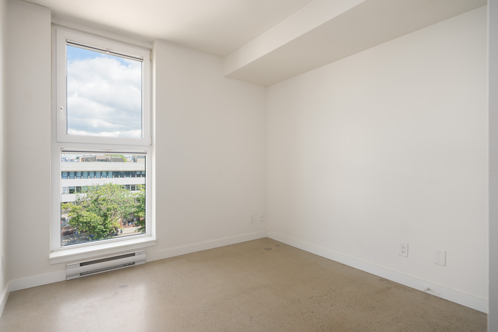 Bright, empty white room with a large window, beige floor, and views of trees and distant buildings outside.