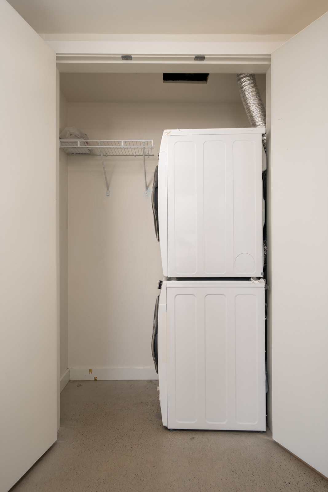 Stacked washer and dryer in a small closet with double doors, left-side wire shelf, exposed vent above, bare floor below.