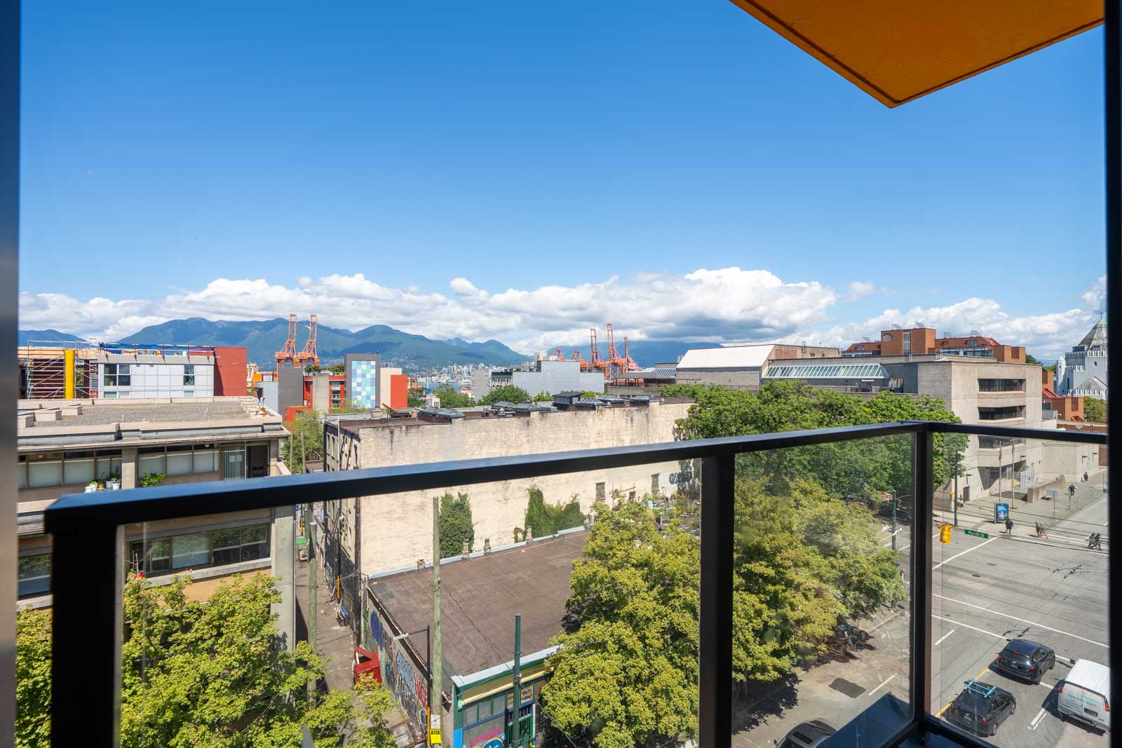 Balcony view of city buildings, leafy trees, a street, and distant mountains under a blue sky with scattered clouds.