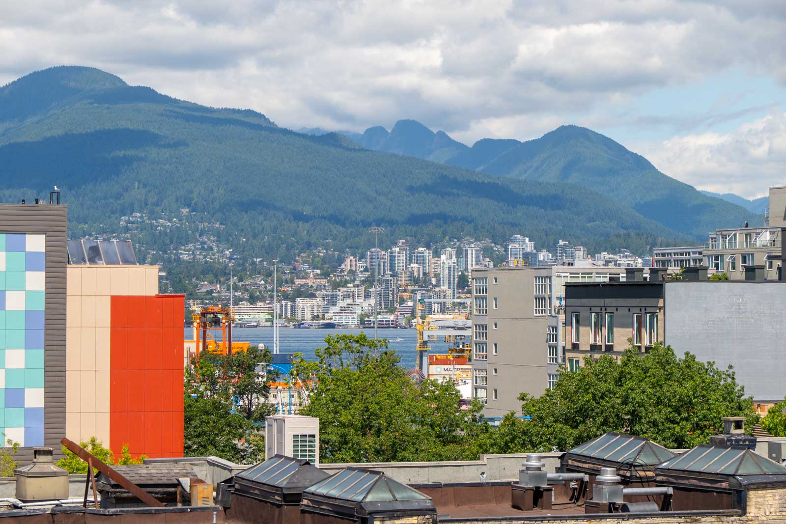 City buildings and trees in the foreground, a body of water, and mountains beneath a cloudy sky in the background.