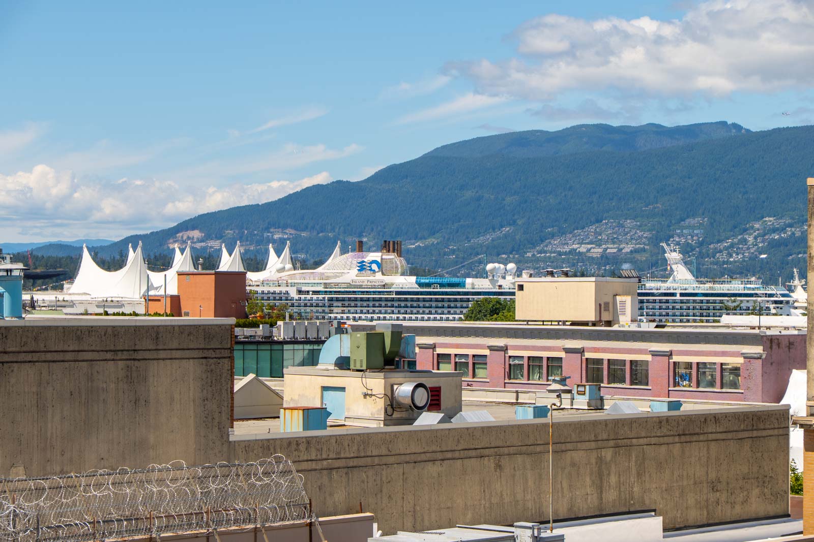 City buildings in front, a large cruise ship at the terminal, and forested mountains behind under a partly cloudy sky.