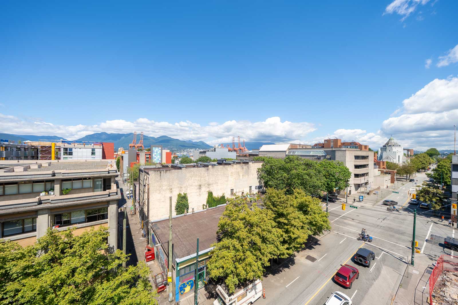 Cars, buildings, and trees at a city intersection beneath a clear blue sky; distant mountains and industrial sites in the background.