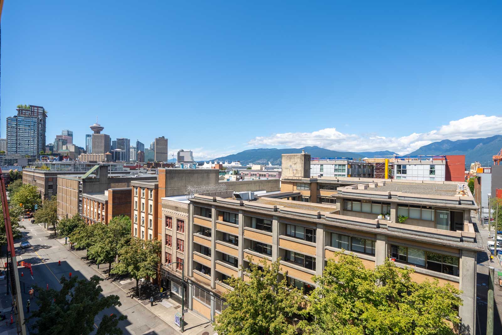City street lined with mid-rise buildings and green trees, distant skyline visible beneath a clear blue sky and mountain backdrop.