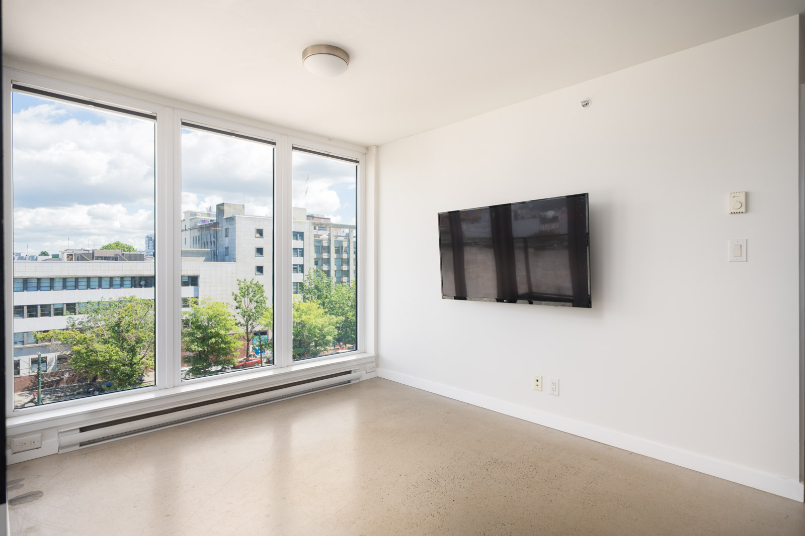 Empty room featuring floor-to-ceiling windows, a wall-mounted TV, plain walls, and views of nearby buildings and trees.