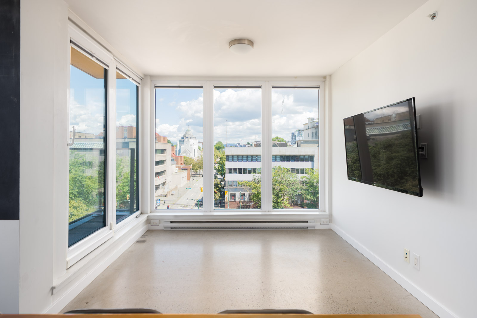 Sunny, unfurnished room with large windows showing city view, white walls, beige floor, and TV mounted on the right wall.