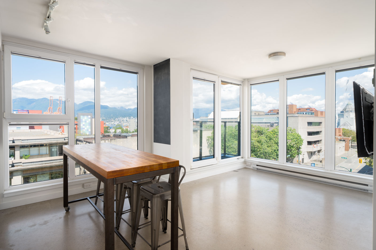 Sunlit room with floor-to-ceiling windows, city and mountain views, a wooden table, and two metal stools on pale flooring.
