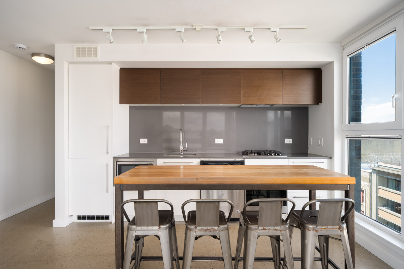 Modern kitchen featuring a wooden island, four metal stools, white and brown cabinets, gray backsplash, sink, stove, and large window.