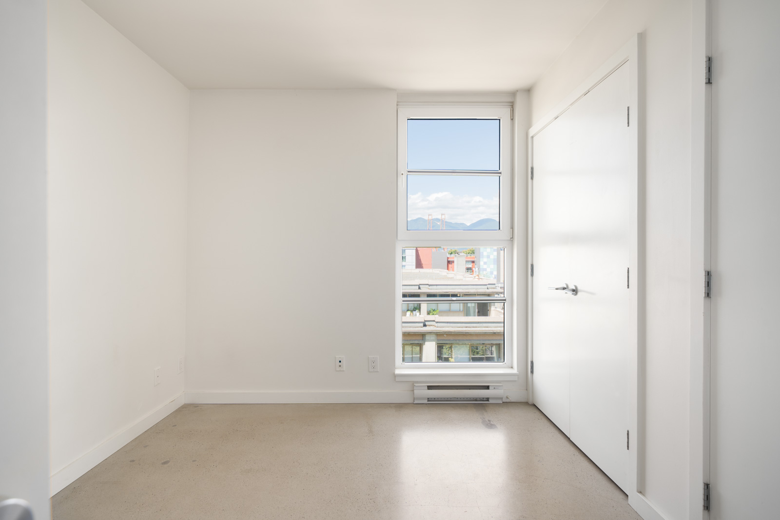Unfurnished white room with a window overlooking buildings and mountains, closed white door on the right, bare floor.