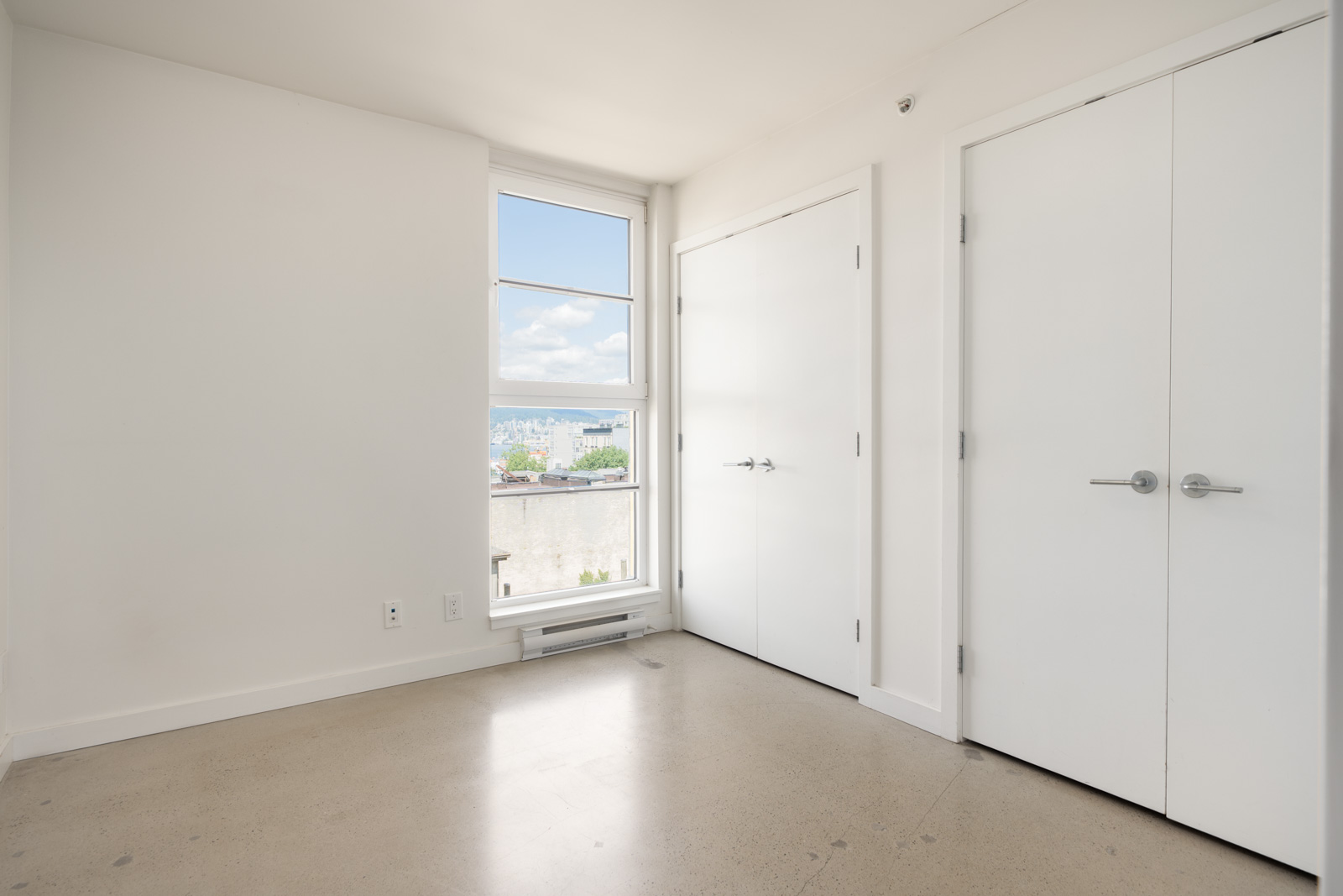 White room with two closed double doors, a large window revealing city buildings and blue sky, white walls, light floor.