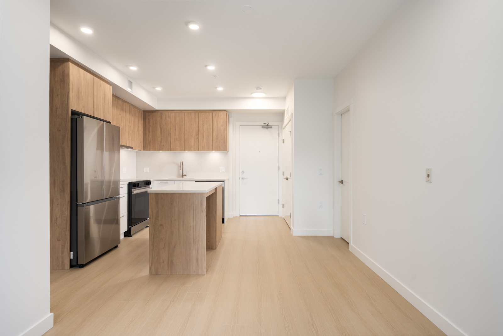 Modern kitchen with light wood cabinets, stainless steel appliances, island, and white walls, open to an area with wood floors and closed door.