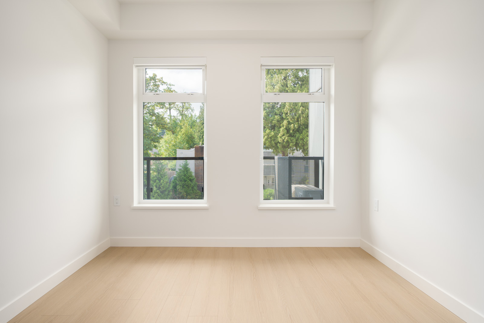 Unfurnished room with light wood floors, white walls, and two large windows facing trees and a balcony railing.