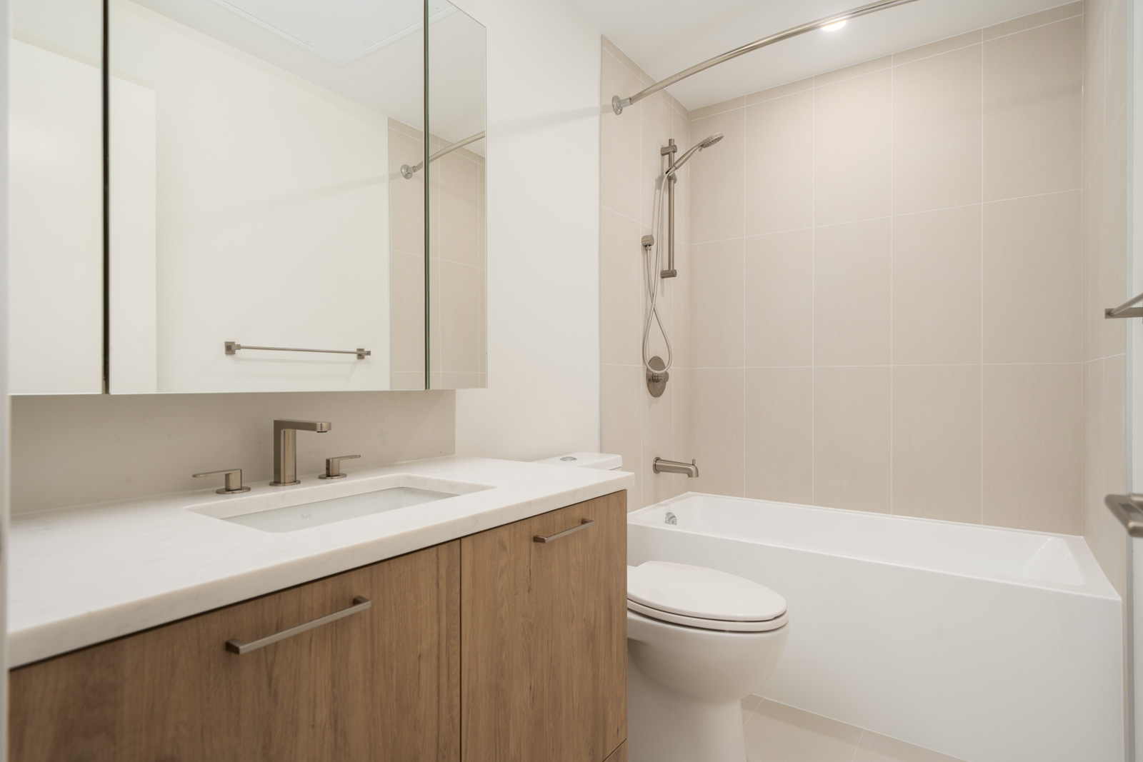 Modern bathroom featuring a wooden vanity, white countertop, built-in sink, mirror cabinet, toilet, and tub-shower with neutral tiles.