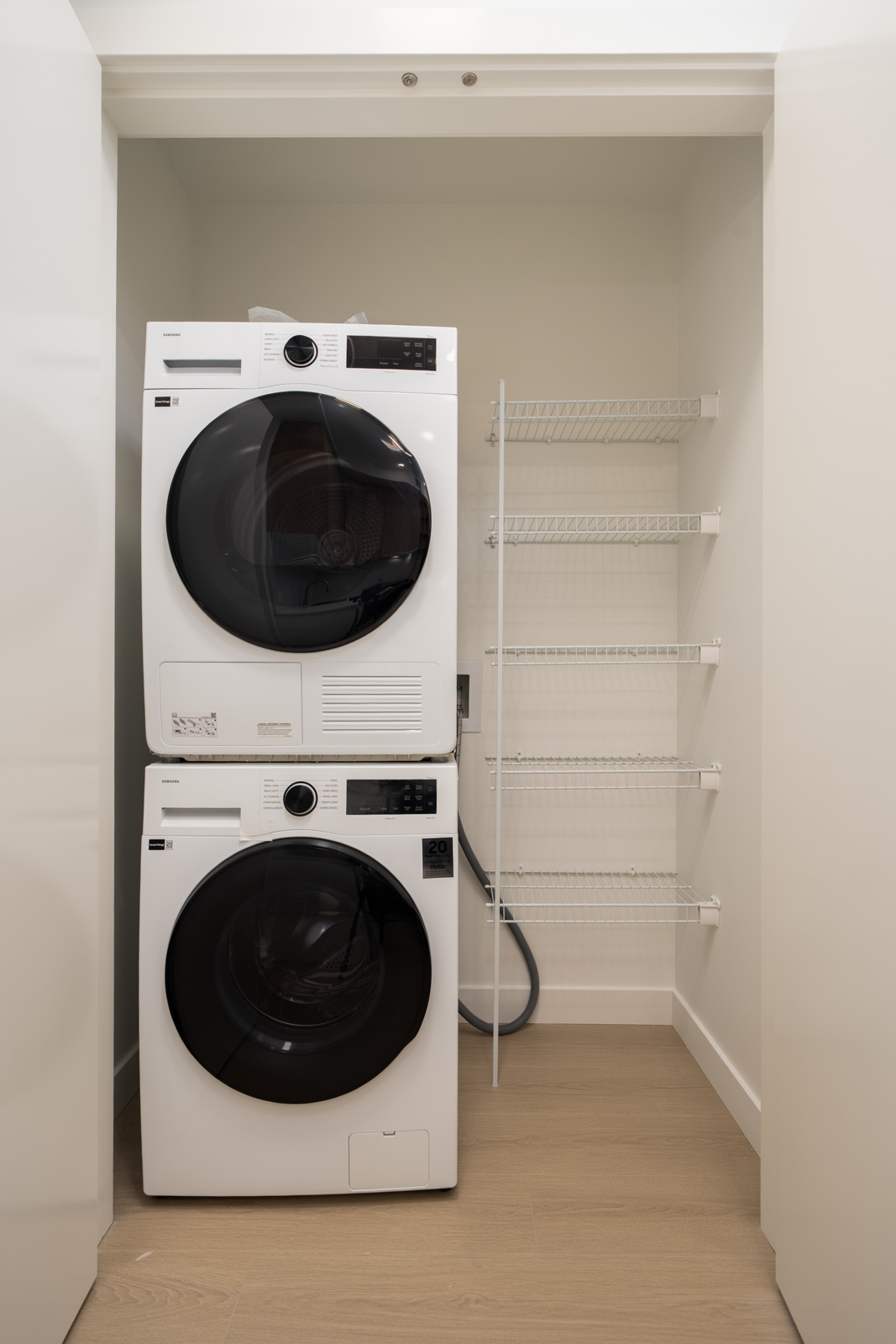 Stacked washer and dryer beside a metal shelf in a compact laundry closet with light wood floors.